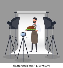 Bloguera culinaria. Un hombre en una plataforma de cocina sostiene un pollo frito en una bandeja. Cámara en un trípode, softbox. Concepto culinario de blog o vlog. Vector.