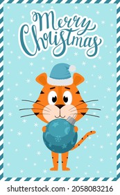 A cute cartoon tiger in a Santa Claus hat stands and holds a round blue Christmas tree toy. Greeting card with hand lettering Merry Christmas. Vector illustration on a blue background with snowflakes