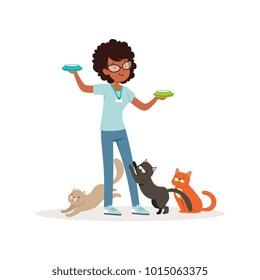 Cute curly-haired girl feeding homeless cats. Volunteer holding bowls with milk. Social help. Teen in glasses, blue t-shirt and jeans. Flat vector design
