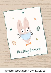 Cute funny Easter Bunny wears a face mask. Healthy Easter handwritten in plain handwriting. Painted Easter eggs and stars around. Greeting card on a wooden table. Easter during the covid-19 pandemic.