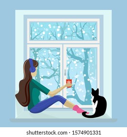 Cute girl in headphones with a cat sitting by the winter window.