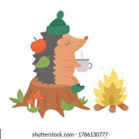 Cute hedgehog sitting on the stump with hot tea near the fire. Vector autumn character isolated on white background. Fall season woodland animal icon.  Funny forest illustration.
