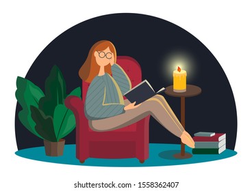 Cute lady in glasses sitting in comfy armchair and reading book. Adorable young woman spending weekend at home. Leisure activity, repose and relaxation. Candle lights the room at night.