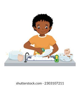 Cute little African boy washing dishes standing at sink in the kitchen doing housework chores at home