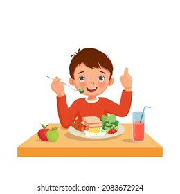 Cute little boy eating breakfast with bread, fried egg, broccoli, vegetables holding fork with sausage showing thumb up gesture.

