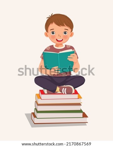 cute little boy student sitting cross legged on stack of books reading
