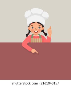 cute little girl in chef hat and apron standing behind empty board showing thumb up and pointing finger to empty banner for copy space