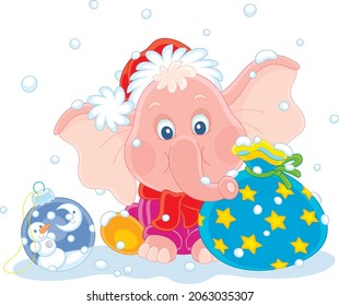 Cute little pink elephant in a red and white Santa hat sitting on snow with a beautiful gift bag and a Christmas toy ball, vector cartoon illustration isolated on a white background