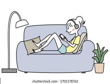 Cute teen girl with headphones relaxing on sofa, using phone. Happy young woman with headphones relaxing on couch, looking at mobile phone. Smiling girl with headphones playing game on smartphone.