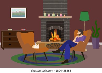 A dark-haired man with a beard in blue trousers and a purple T-shirt sits in an orange chair in a room with a burning dark brick fireplace, opposite a three-colored cat sits in another chair