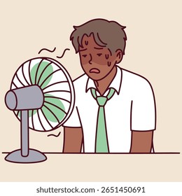 Dark-skinned man leans on table near small fan, visibly tired and sweating. He suffers from summer heat in indoor space