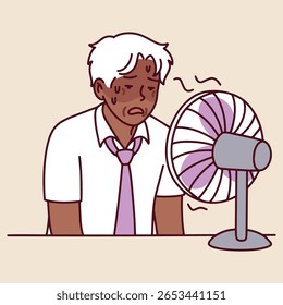 Sênior de pele escura senta-se à mesa ao lado do ventilador e limpa o suor. Ele luta com altas temperaturas em ambientes fechados