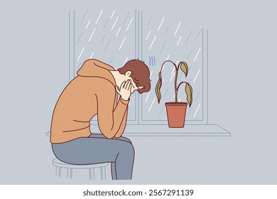 Depressed man sits alone and suffers from melancholy in room with window and wilted flower. Melancholy in older guy victim of autumn blues or everyday problems and troubles at work