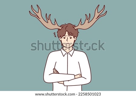 Disgruntled guy with deer horns on head stands with arms crossed and looks at screen with displeasure. Metaphor of man who suffered from infidelity of girlfriend or adultery wife