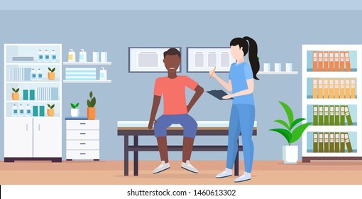 doctor holding clipboard female therapist consulting injured african american male patient sitting on bed manual sport physical therapy concept modern medical office interior horizontal