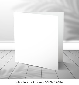A double blank closed card stands near a white wall with a shadow of a palm tree on plank floor. Mockup for design