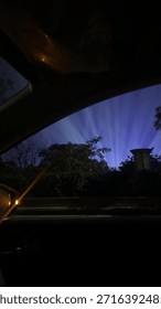 Dramáticos rayos de luz azul iluminan el cielo nocturno sobre los árboles y el entorno urbano, visto desde el interior de un automóvil a través de una ventana. El oscuro primer plano y el brillante cielo crean una atmósfera animada y cinematográfica, que representa la vida nocturna, los eventos de la ciudad, la iluminación al aire libre, el misterio, los viajes y el ambiente urbano moderno. Adecuado para fondos creativos, conceptos abstractos, imágenes de viajes nocturnos y uso de diseño atmosférico.