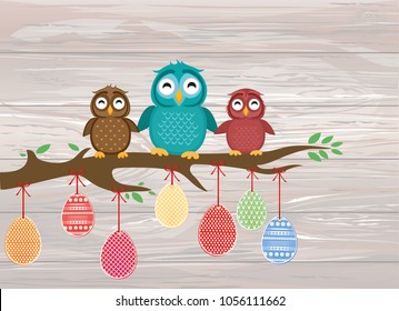 Easter eggs hang on ribbons. Lovely owls are sitting on a tree branch. Greeting card or invitation for a holiday. Vector on wooden background.