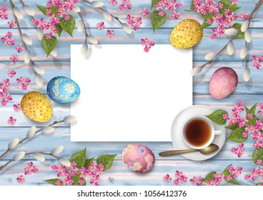 Easter holiday background with a cup of tea, flowers, painted eggs, and a blank piece of paper on wooden background. Top view composition