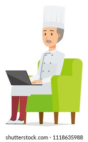 An elderly male chef wearing a cook coat is sitting on a sofa and operating a laptop computer