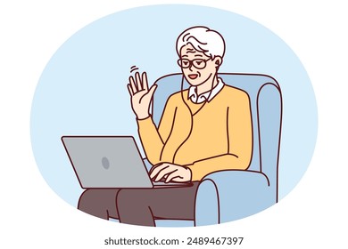 Elderly man makes video call through laptop sitting in chair and waving hand greeting interlocutor. Elderly human with laptop learns computer literacy by participating in online webinar