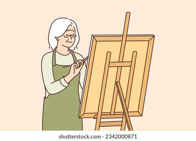 Elderly woman artist stands near easel and draws picture enjoying creative hobby after retiring. Gray-haired grandmother is fond of drawing and dreams of becoming famous artist or selling own artworks