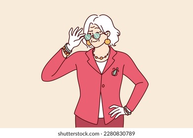 Elderly woman in elegant suit for going to work or dinner party adjusts glasses in front of eyes. Gray-haired grandmother of retirement age dressed in fashionable elegant clothes looks at camera 