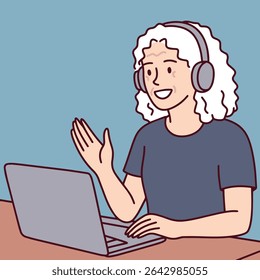 Elderly woman with gray hair wears headphones and waves while using laptop for remote communication and online video call from home