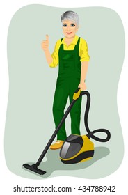 Elderly woman in green coveralls posing with vacuum cleaner and giving thumbs up