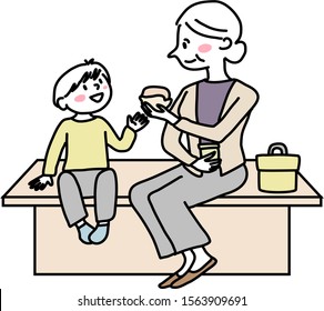 Elderly woman and her grandchild sitting together on bench, have lunch. Grandma and grandson sitting on bench and eating sandwiches. Grandmother and little boy eating food together outdoors.