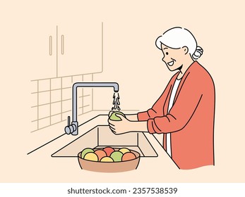 Elderly woman washes fruits in sink, standing in kitchen and observing rules of hygiene to avoid infection. Grandmother washes vegetables before preparing dinner, following recommendations of doctors