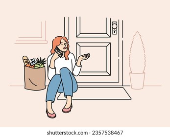 Embarrassed woman sits on threshold near door with jammed lock and cannot get into house, makes phone call. Puzzled girl with bag of groceries, calling specialist because of loss of key to door