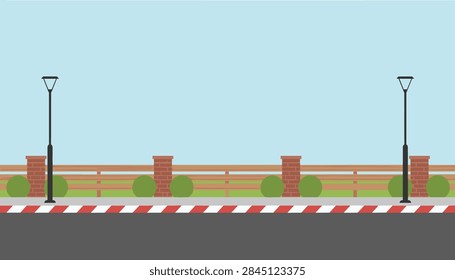Empty asphalt road bordered by a classic wooden fence, green bushes, and two street lamps under a clear blue sky.
