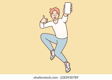 Encouraged man jumps up with mobile phone in hands showing blank screen of gadget and shows thumbs up. Excited teenager guy with smartphone advertises mobile app or website for young people 