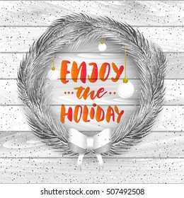 Enjoy the holiday - freehand hand drawn calligraphic design for Xmas greetings cards, invitations. Christmas hand drawn lettering in pine branches wreath on white wooden background.