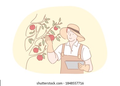 Farming, agriculture, harvesting concept. Smiling young man farmer in working apron, gloves and hat standing with documents and touching fresh red ripe apples on tree in farmland for harvesting 