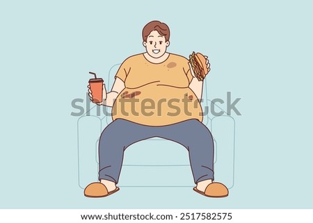 Fat man suffers from obesity caused by problem of overeating and smiles, sitting on sofa with burger and soda in hands. Fat guy eats junk food due to lack of willpower or genetic disease