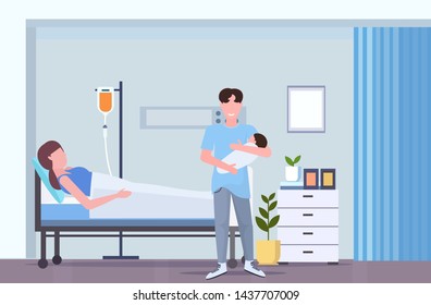 father holding newborn baby man visiting his wife lying in hospital bed with dropper happy family parenthood childbirth concept maternity ward interior flat full length horizontal