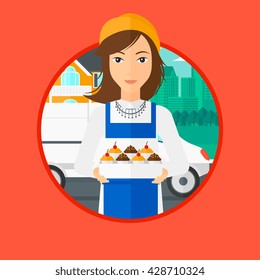 A female baker holding a box of cakes. A baker delivering cakes. Baker with cupcakes standing on the background of delivery truck. Vector flat design illustration in the circle isolated on background.