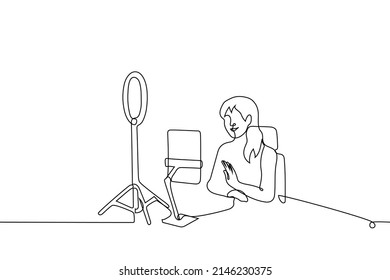 female blogger smiles and waves at the camera of tablet on tripod, it is illuminated by ring lamp for video light - one line drawing vector. concept of online streaming, recording video lesson