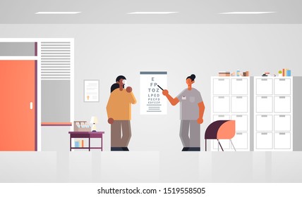 female doctor ophthalmologist pointing at letters on eye chart checking african american patient vision medicine healthcare concept hospital medical clinic office interior full length horizontal