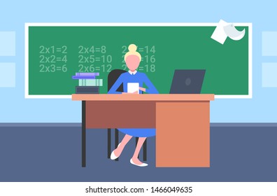 female teacher sitting at desk in front of chalkboard using laptop. math lesson education concept. modern school classroom interior horizontal flat.