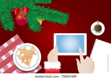 Finger of Santa Claus is touching a blank screen of tablet ready for your text over the table with  branch of Christmas tree, Christmas sweets on the plate and  cup of coffee.