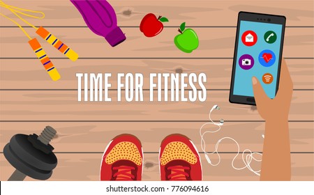 Fitness, healthy and active lifestyles concept. Dumbbells, sport shoes, skipping rope, smart phone with earphone and bottle on wooden gym floor with word time for fitness. Hand holding smartphone.