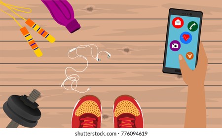 Fitness, healthy and active lifestyles concept. Dumbbells, sport shoes, skipping rope, smart phone with earphone and water bottle on wooden gym floor. Hand holding smartphone with icons