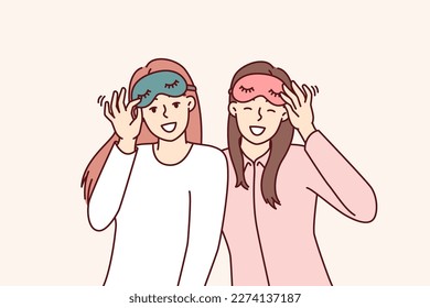 Friendly girls in pajamas with sleep masks are preparing to relax in comfortable bedroom with soft bed. Happy women use eye masks to protect themselves from sunlight during daytime naps or siestas 