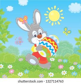 Friendly smiling Easter Bunny with a colorfully decorated egg walking among flowers on green grass on sunny spring day, vector illustration in a cartoon style