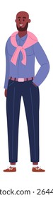 Full length portrait of a dark skinned man with a beard and bald head, wearing a pink scarf, blue shirt and dark blue pants, smiling with one hand in his pocket