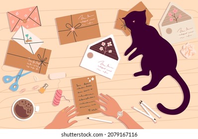 Funny cat lying and watching the woman's hands on the table. Top view of the desk with a sheet of paper, letters, cat, scissors, coffee, notepad, crumpled paper balls and supplies.