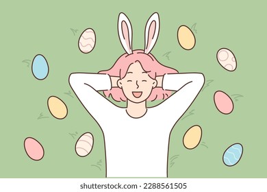 Funny girl with rabbit ears lies on grass among colorful Easter eggs and smiles broadly. Carefree female teenager rejoice at onset of traditional Christian holiday of Easter Sunday